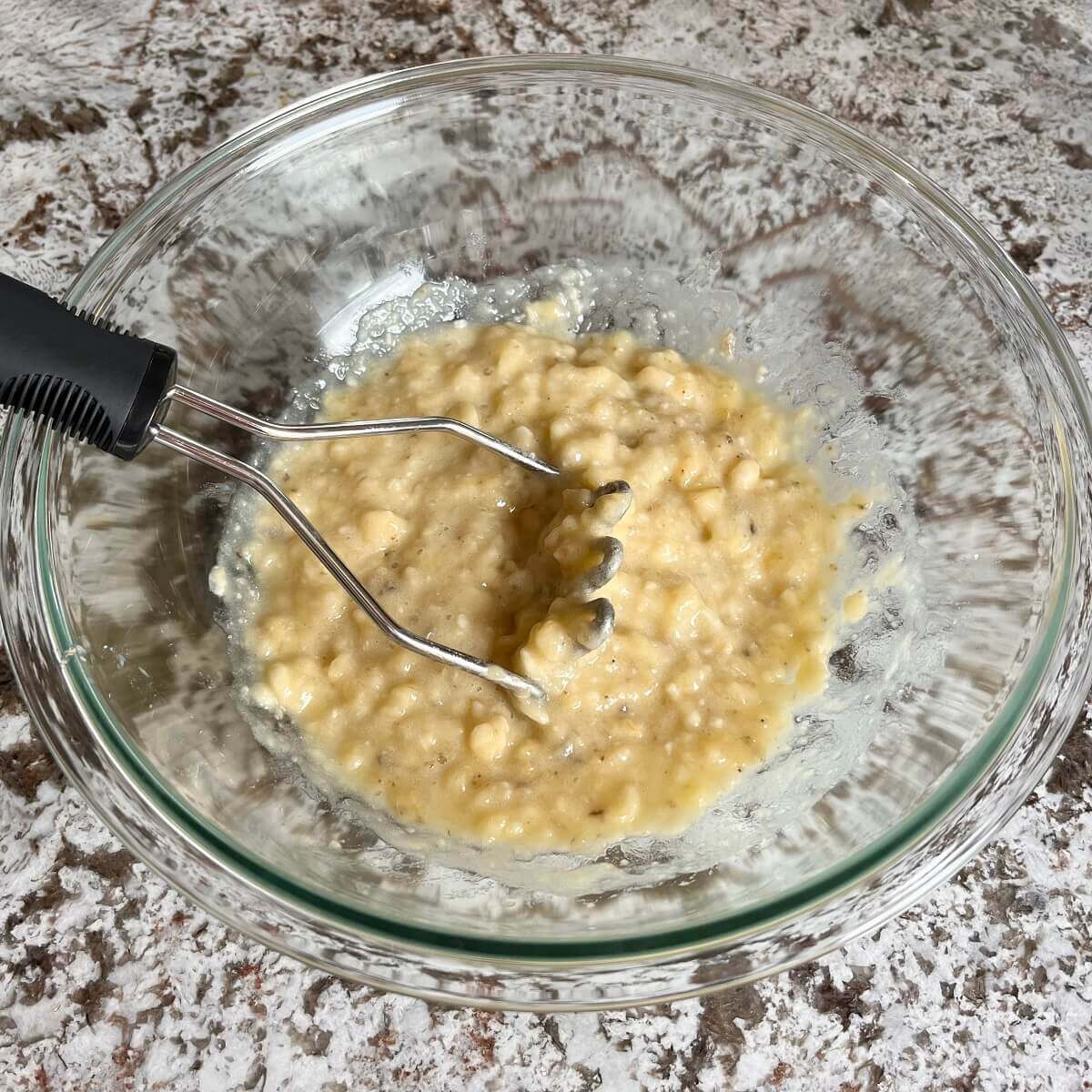 Mashed bananas in a glass bowl with a potato masher.